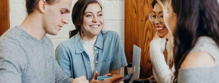 Ausgezeichnet.org thematisiert Interaktives Marketing und nennt die Vorteile eines stetigen Austausch mit den Kunden, das Bild zeigt 4 Menschen, die lachend zusammen an einem Tisch sitzen und an verschiedenen Geräten arbeiten