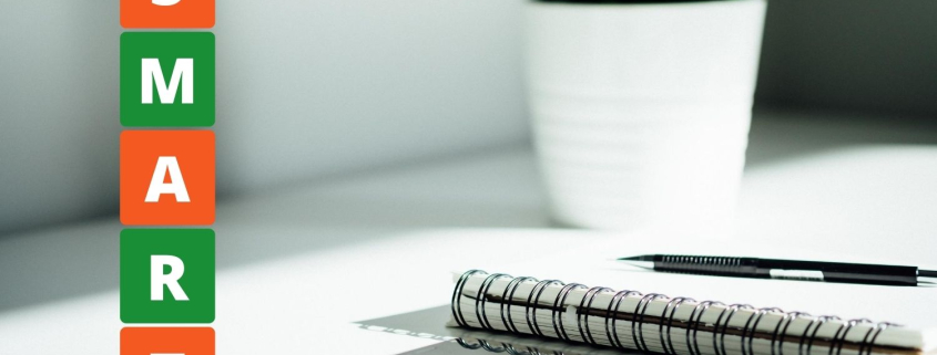 Ausgezeichnet.org erlärt, wie man sich mit der SMART-Methode bessere Ziele setzt und diese auch erreicht, das Bild zeigt ein Notizbuch und Smartphone auf einem Tisch, auf dem Bild ist das Wort SMART abgebildet