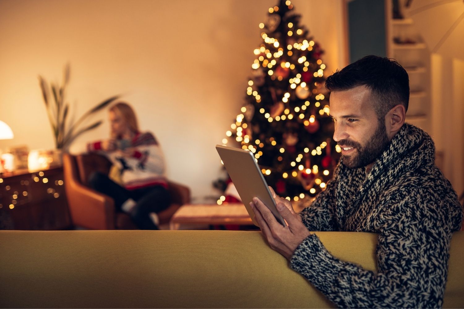 Ein Mann sitzt beim Weihnachtsbaum und hält ein Tablet in den Händen